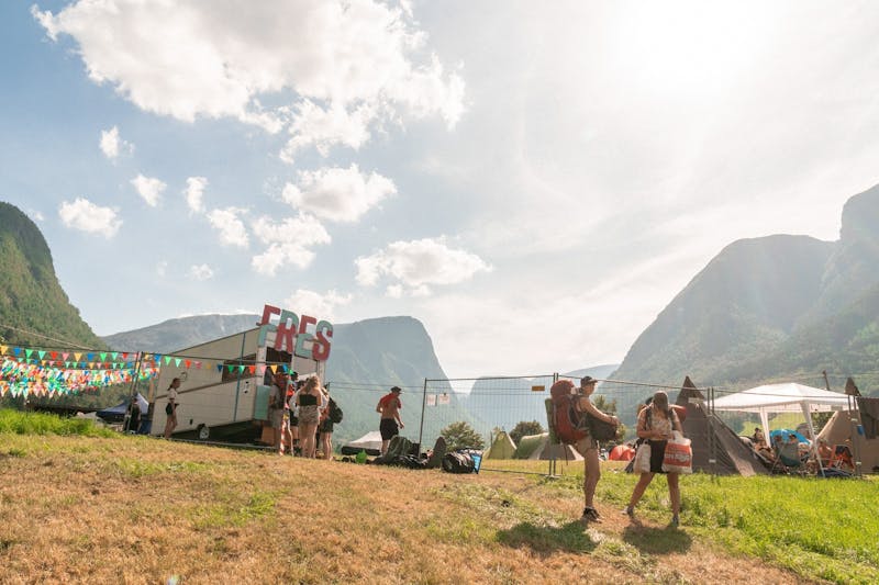 Bildet viser en solfull dag på Fres festival. I forgrunnen står et par campere med sekk og handleposer. I bakgrunnen er det en bås med Fres-logo, bak der er fjell.